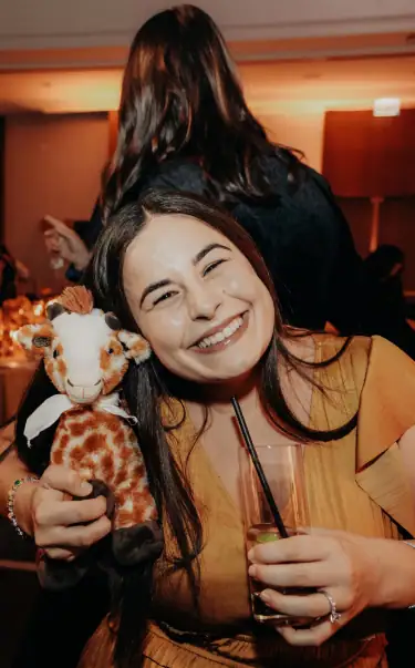 Cute young woman smiling holding a plush giraffe at Camp One Step event, joyful and fun atmosphere.