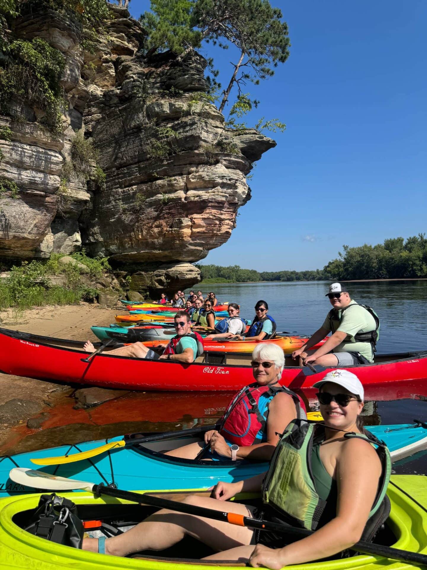 Vibrant group kayaking along scenic river with rocky cliffs and lush greenery at Camp One Step.