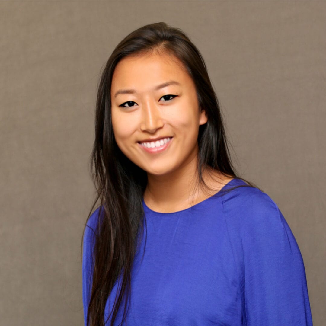 Young woman smiling, vibrant blue top, indoor professional setting, friendly, welcoming atmosphere.