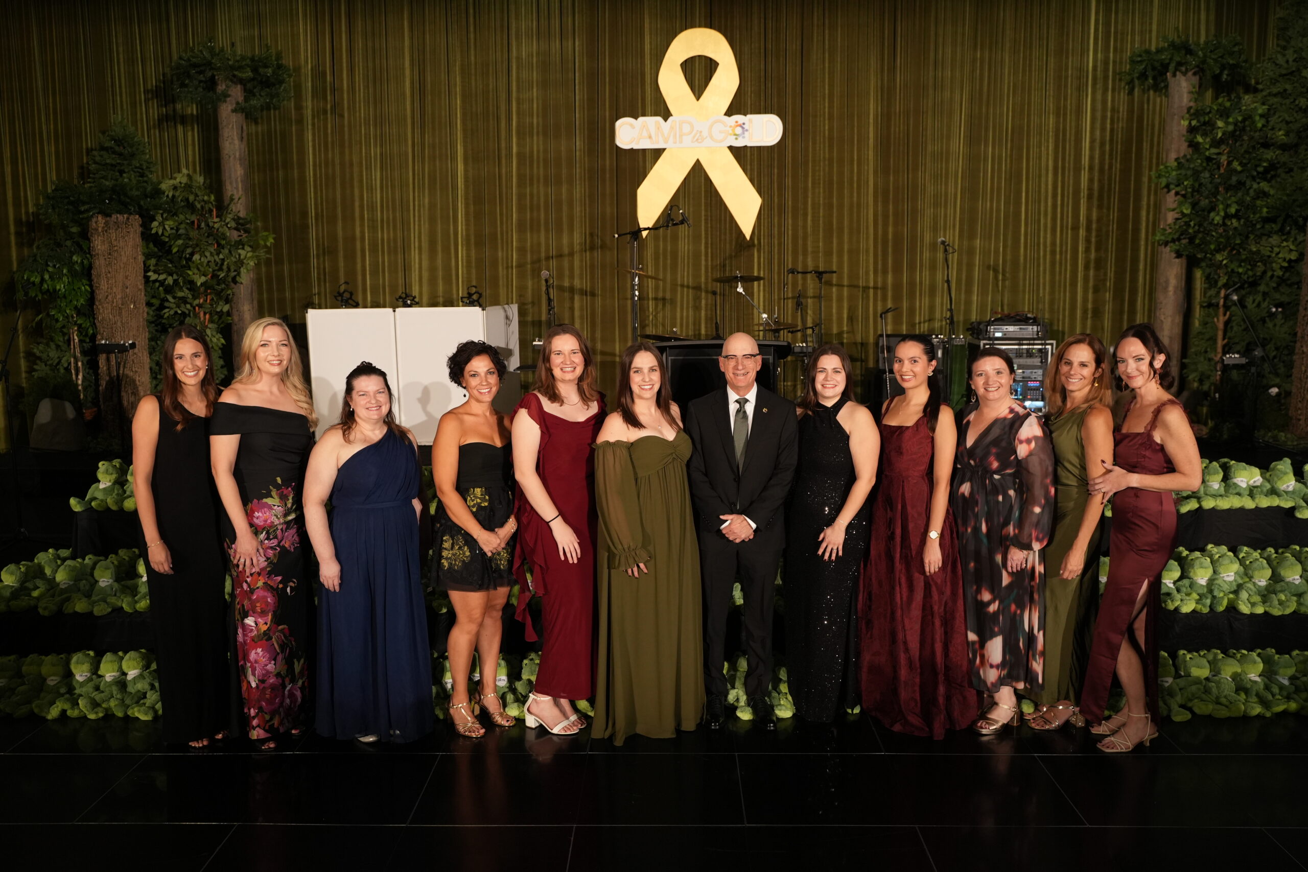 A group of women and one man posing at Camp One Step charity event, with a stage and musical instruments in the background.