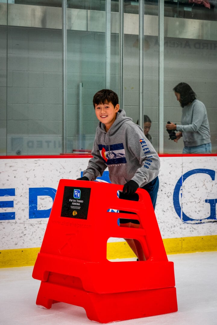 Child smiling wearing gray hoodie holding a red skating aid on ice rink.