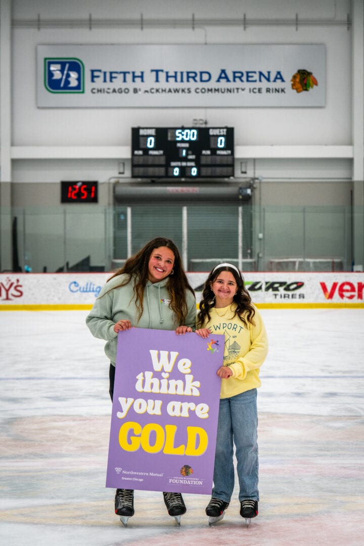 Female youth ice skaters on ice rink holding a "We think you are GOLD" sign, smiling and celebrating.