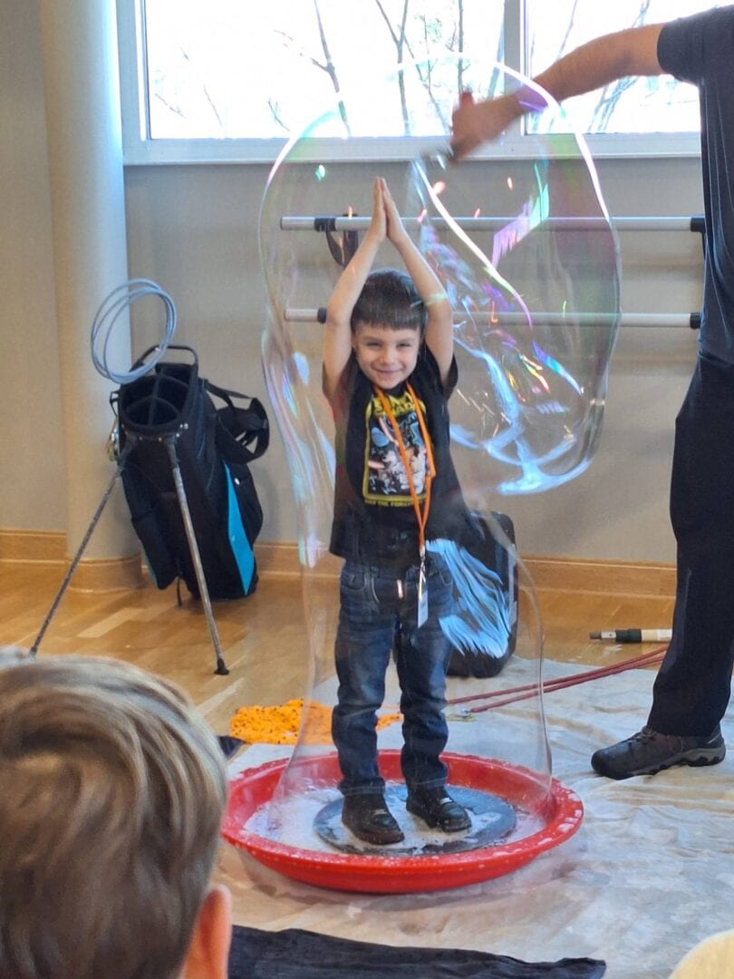 Child inside large soap bubble, kids engaging in science and outdoor fun at camp.