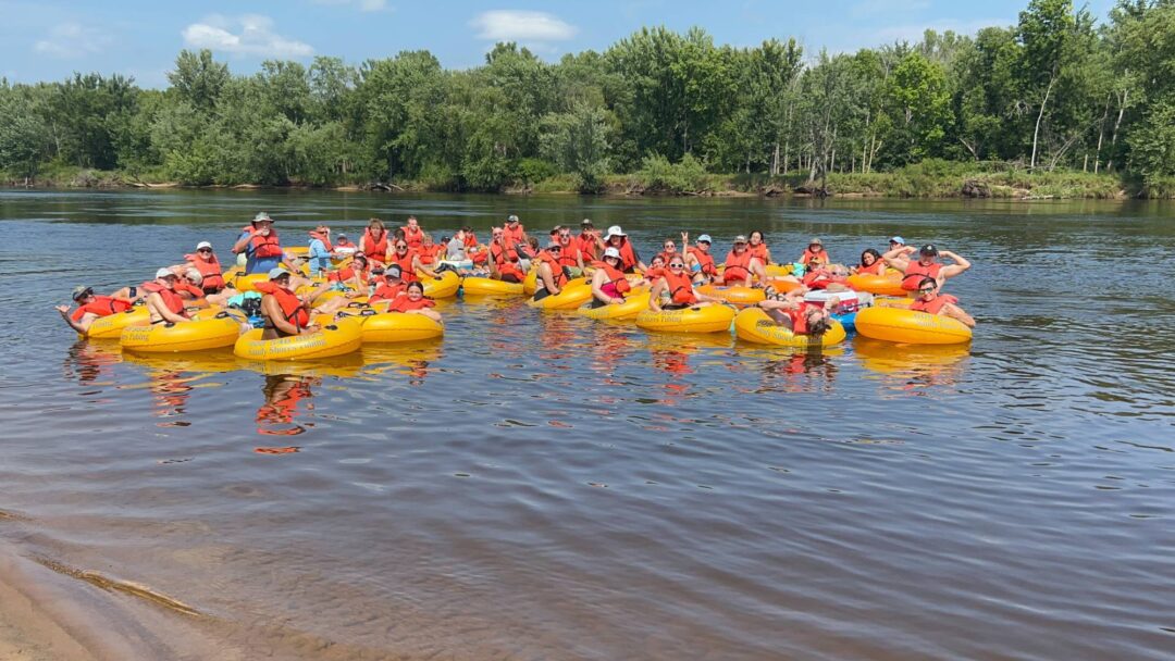 Group of kids and adults enjoying tubing at Camp One Step on the river with lush green trees in the background.