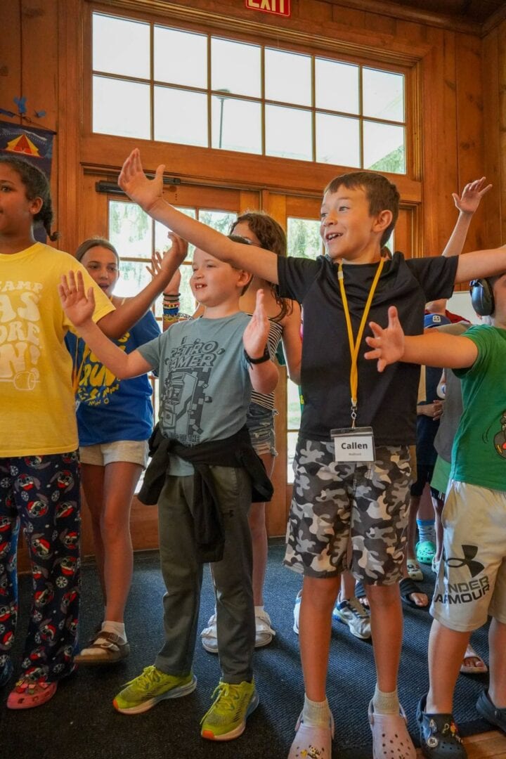 Children smiling and most with arms raised inside a wooden cabin at Camp One Step.