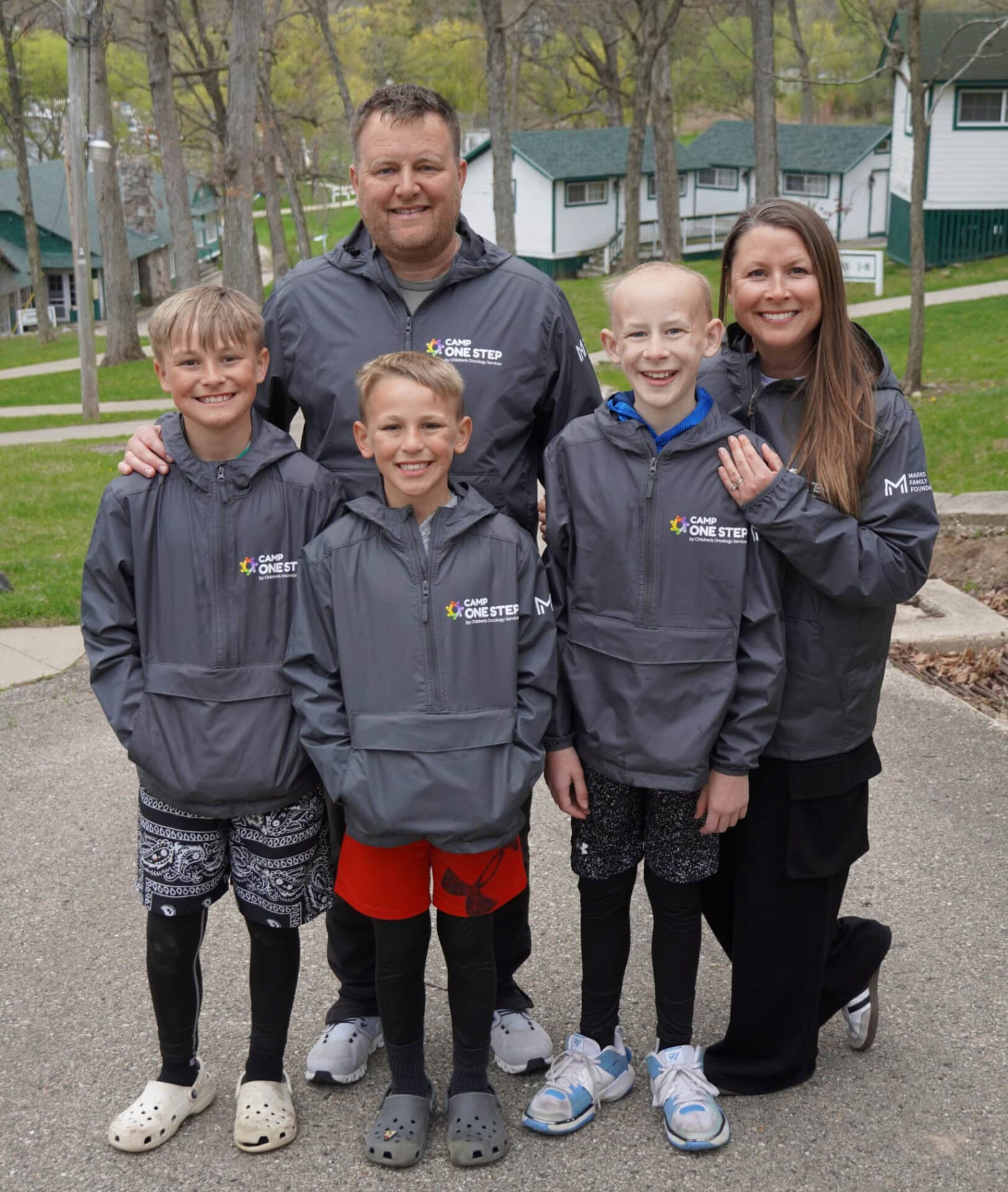 - Diverse family group wearing Camp One Step jackets outdoors in a wooded campground setting.
