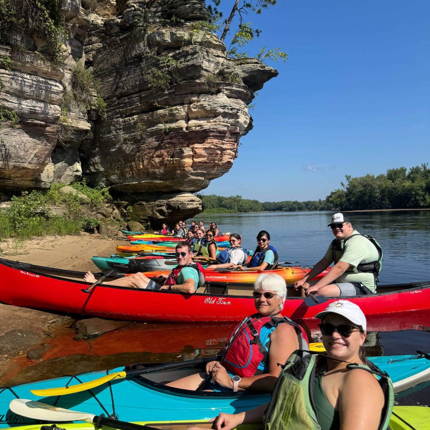 Vibrant group kayaking along scenic river with rocky cliffs and lush greenery at Camp One Step.