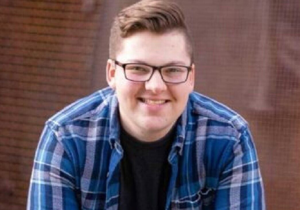 Smiling young man in glasses wearing a blue plaid shirt, outdoor setting.